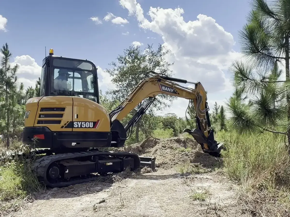 Compact excavator working along a sandy route between young pines.